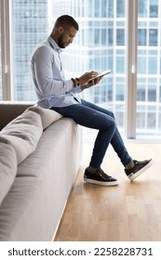 Focused young entrepreneur man using tablet at home for Internet communication, working on job tasks, making payment with bank application, sitting on sofa against big window city view